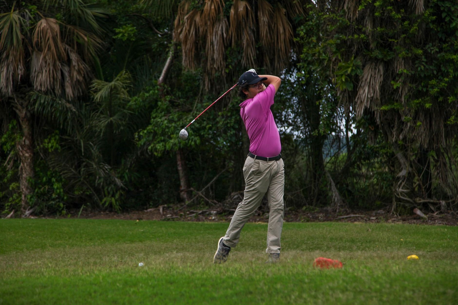 Adult male golfer swinging a club on a lush green golf course surrounded by trees.